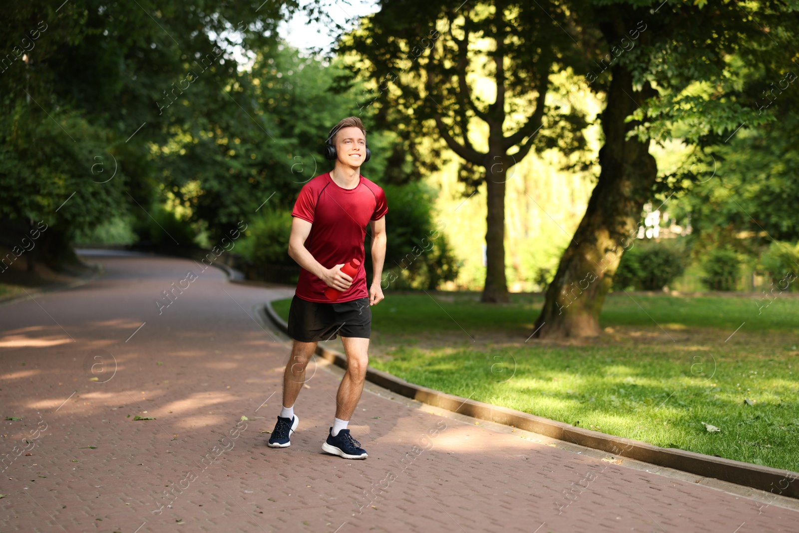 Photo of Smiling sportsman in headphones running with bottle outdoors. Morning exercise