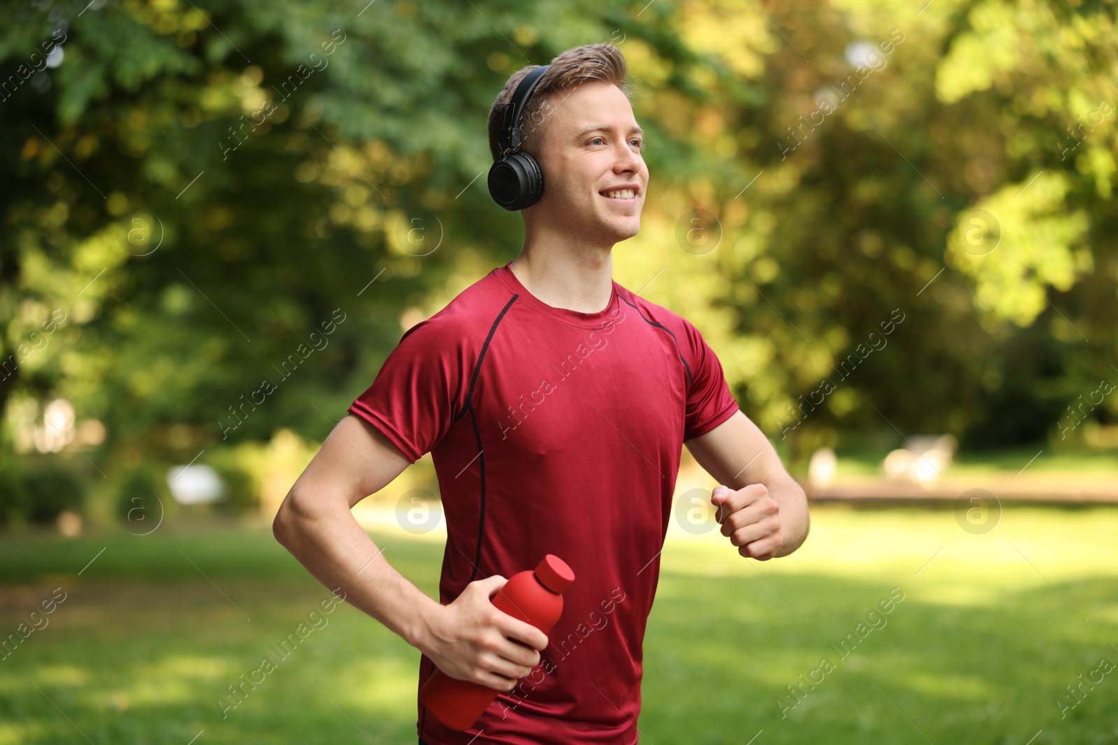 Photo of Smiling sportsman in headphones running with bottle outdoors. Morning exercise