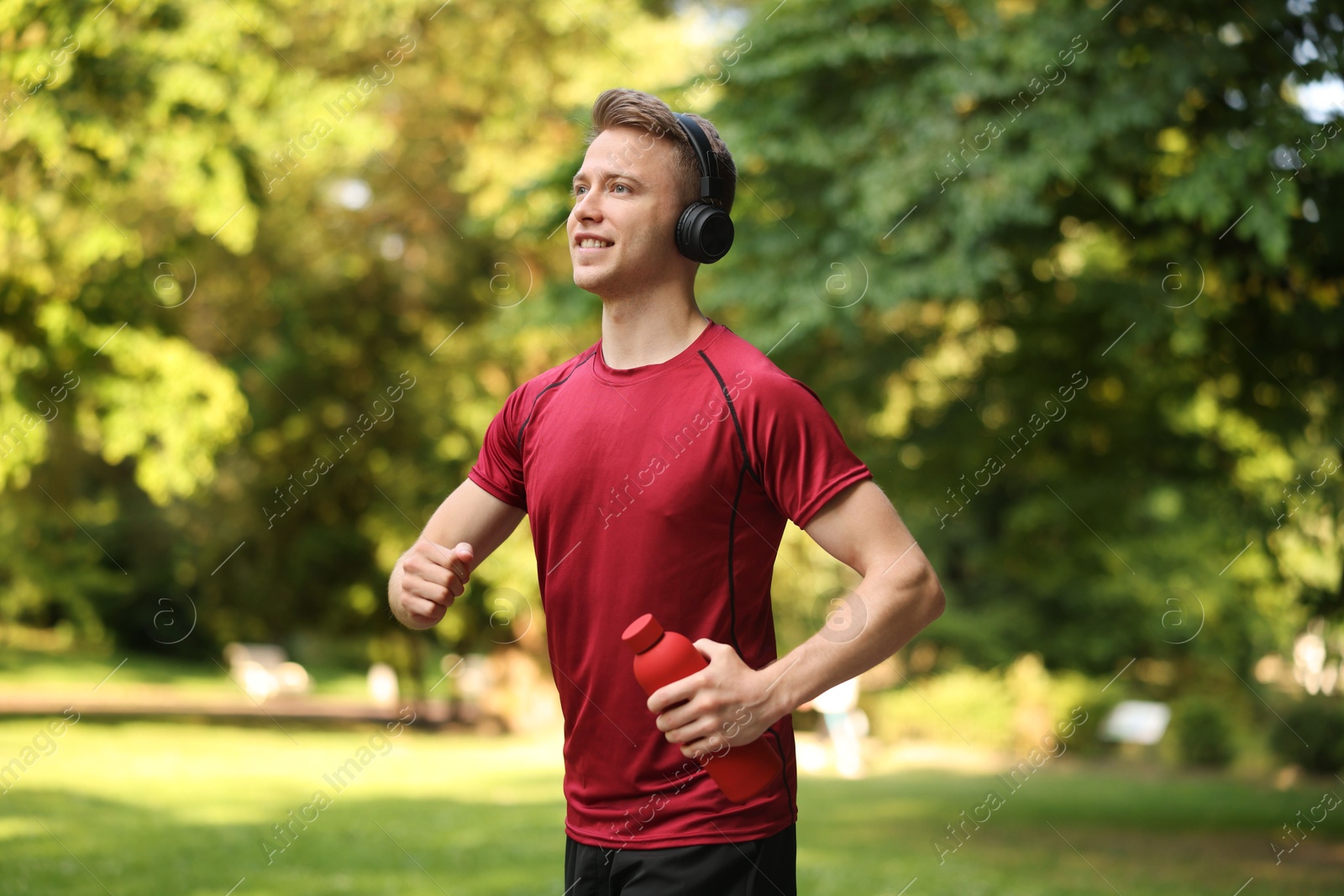 Photo of Smiling sportsman in headphones running with bottle outdoors. Morning exercise