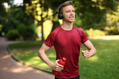 Photo of Smiling sportsman in headphones running with bottle outdoors. Morning exercise