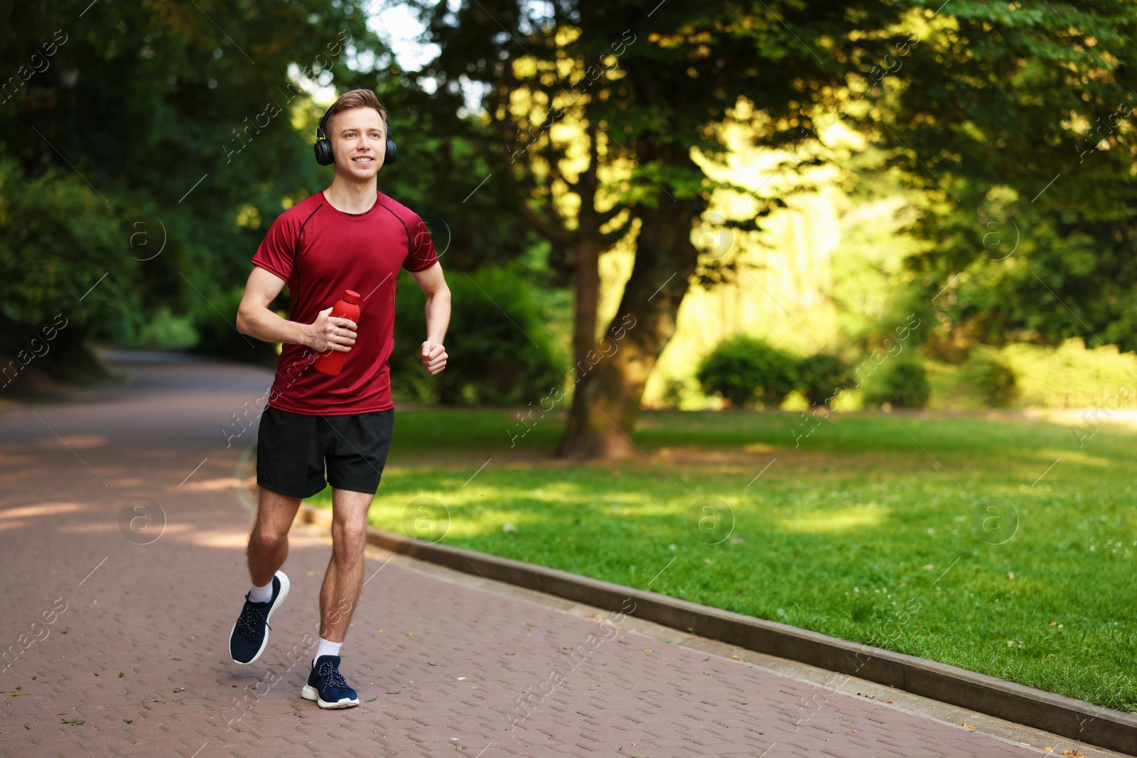 Photo of Smiling sportsman in headphones running with bottle outdoors, space for text. Morning exercise