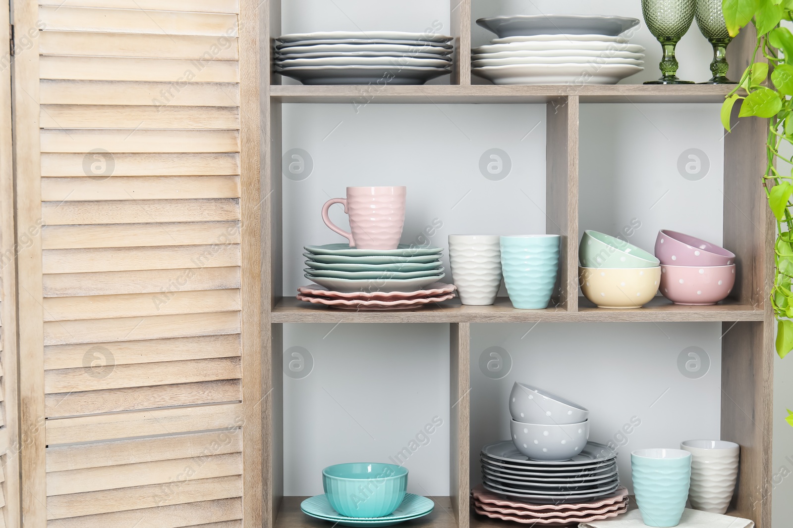 Photo of Shelving unit with different dishware, glasses, cups and floral decor near white wall indoors