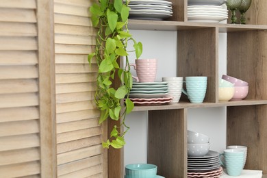 Photo of Shelving unit with different dishware, cups and floral decor near white wall indoors