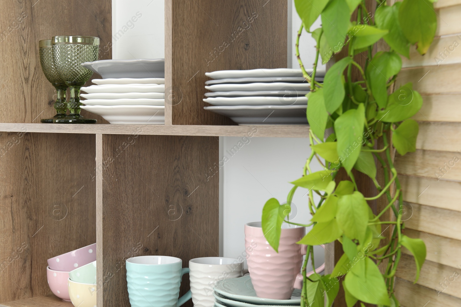 Photo of Shelving unit with different dishware, glasses, cups and floral decor near white wall indoors, closeup