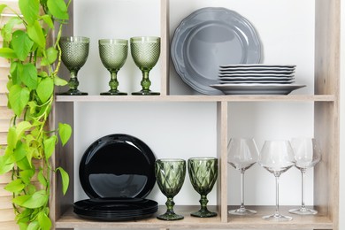 Photo of Shelving unit with different dishware, glasses and floral decor near white wall indoors