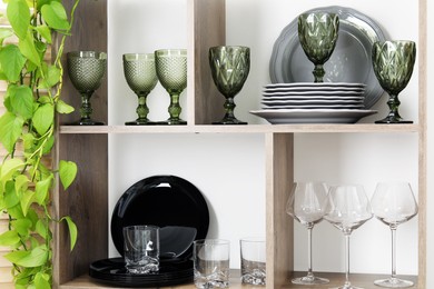Photo of Shelving unit with different dishware, glasses and floral decor near white wall indoors