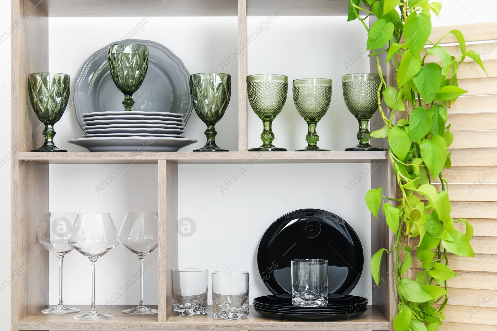 Photo of Shelving unit with different dishware, glasses and floral decor near white wall indoors