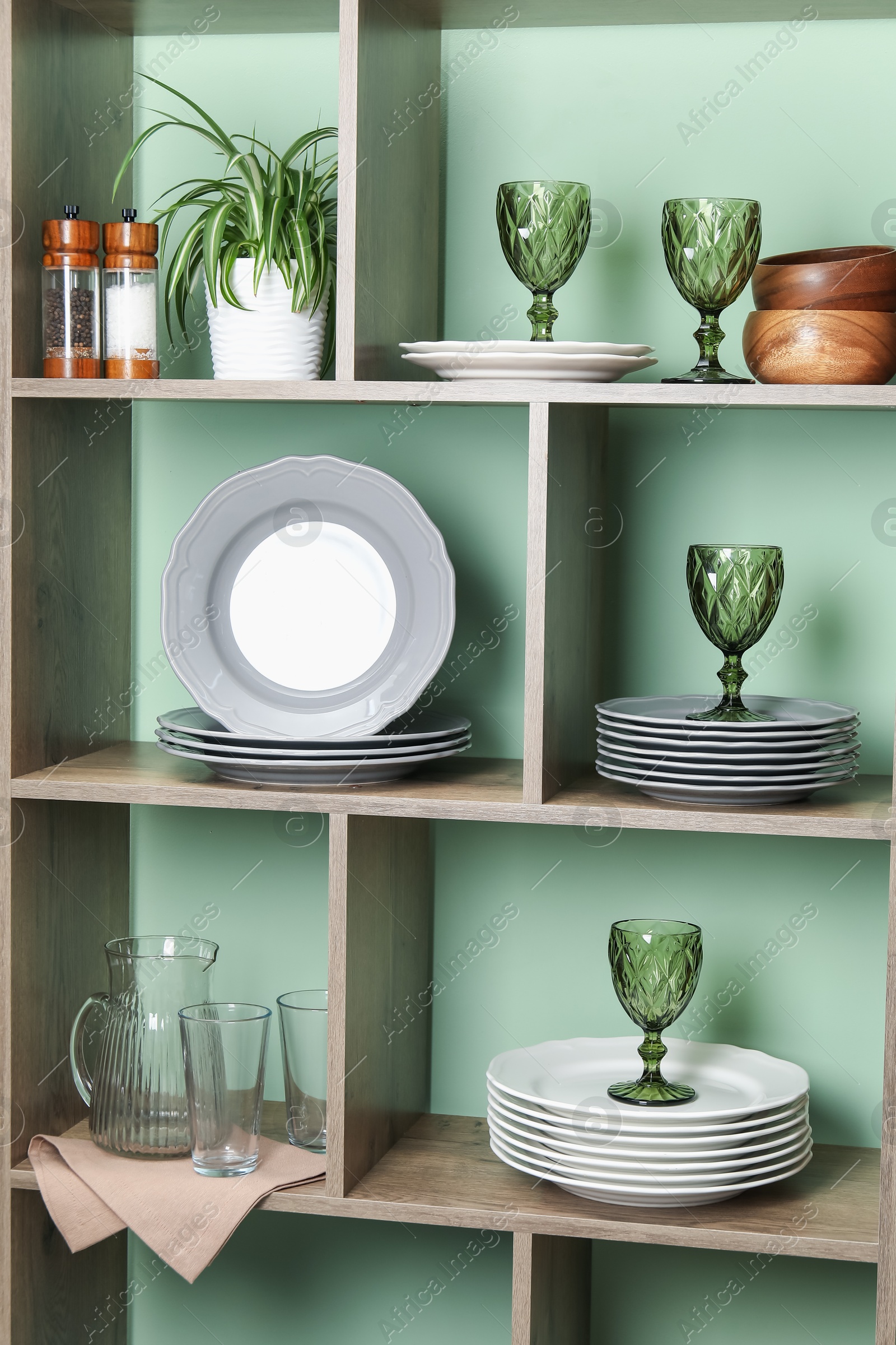 Photo of Shelving unit with different dishware, glasses and floral decor near green wall indoors