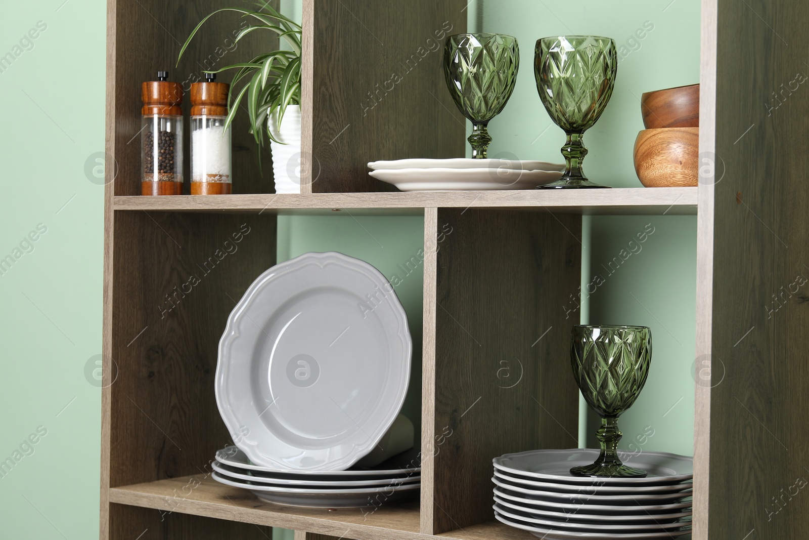 Photo of Shelving unit with different dishware, glasses and floral decor near green wall indoors, closeup