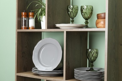 Photo of Shelving unit with different dishware, glasses and floral decor near green wall indoors, closeup