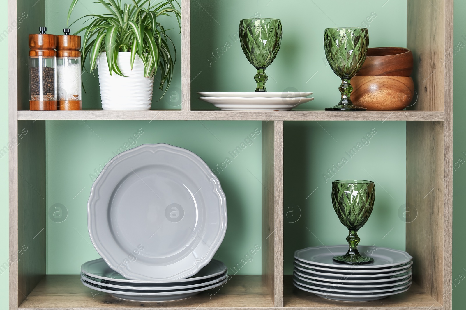 Photo of Shelving unit with different dishware, glasses and floral decor near green wall indoors