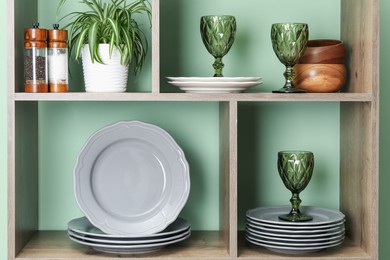 Photo of Shelving unit with different dishware, glasses and floral decor near green wall indoors