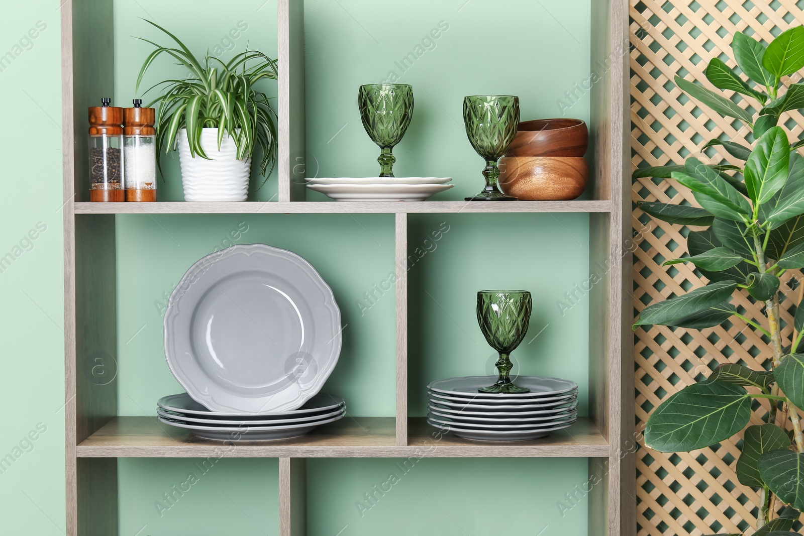 Photo of Shelving unit with different dishware, glasses and floral decor near green wall indoors