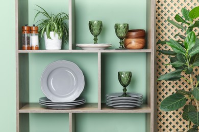 Photo of Shelving unit with different dishware, glasses and floral decor near green wall indoors