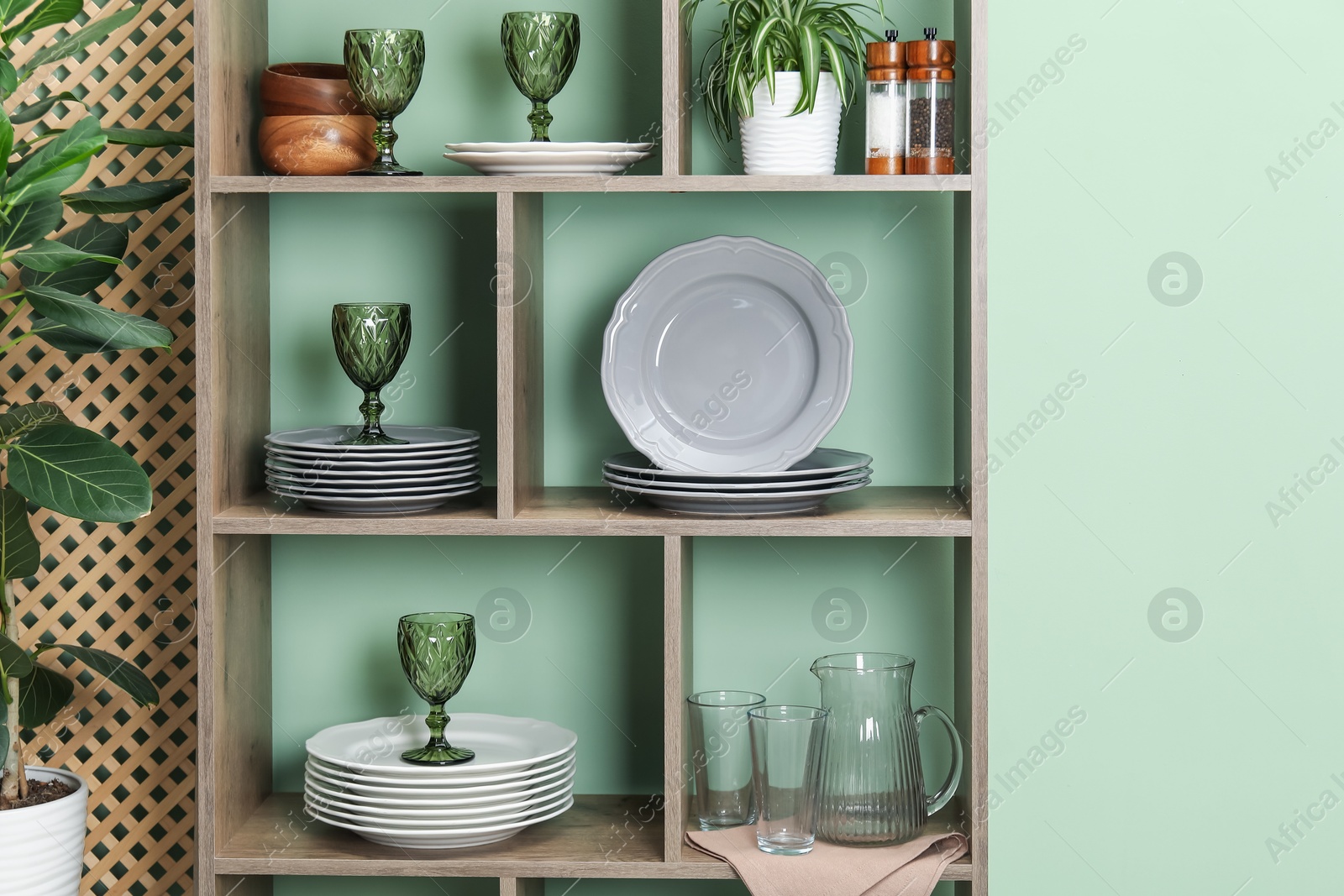 Photo of Shelving unit with different dishware, glasses and floral decor near green wall indoors