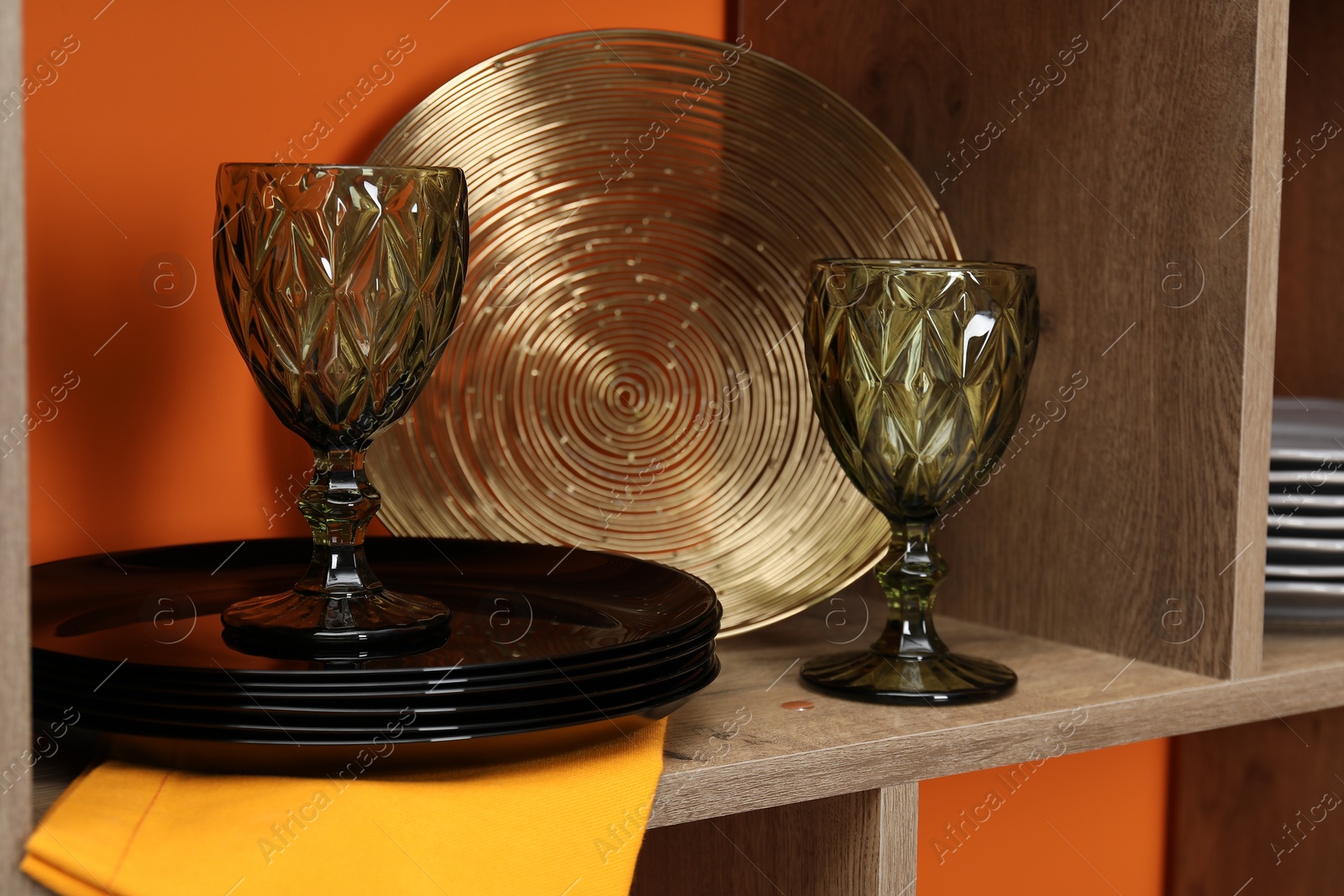 Photo of Different dishware and glasses on wooden shelf near orange wall indoors, closeup