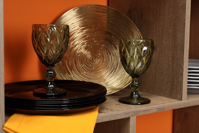 Photo of Different dishware and glasses on wooden shelf near orange wall indoors, closeup