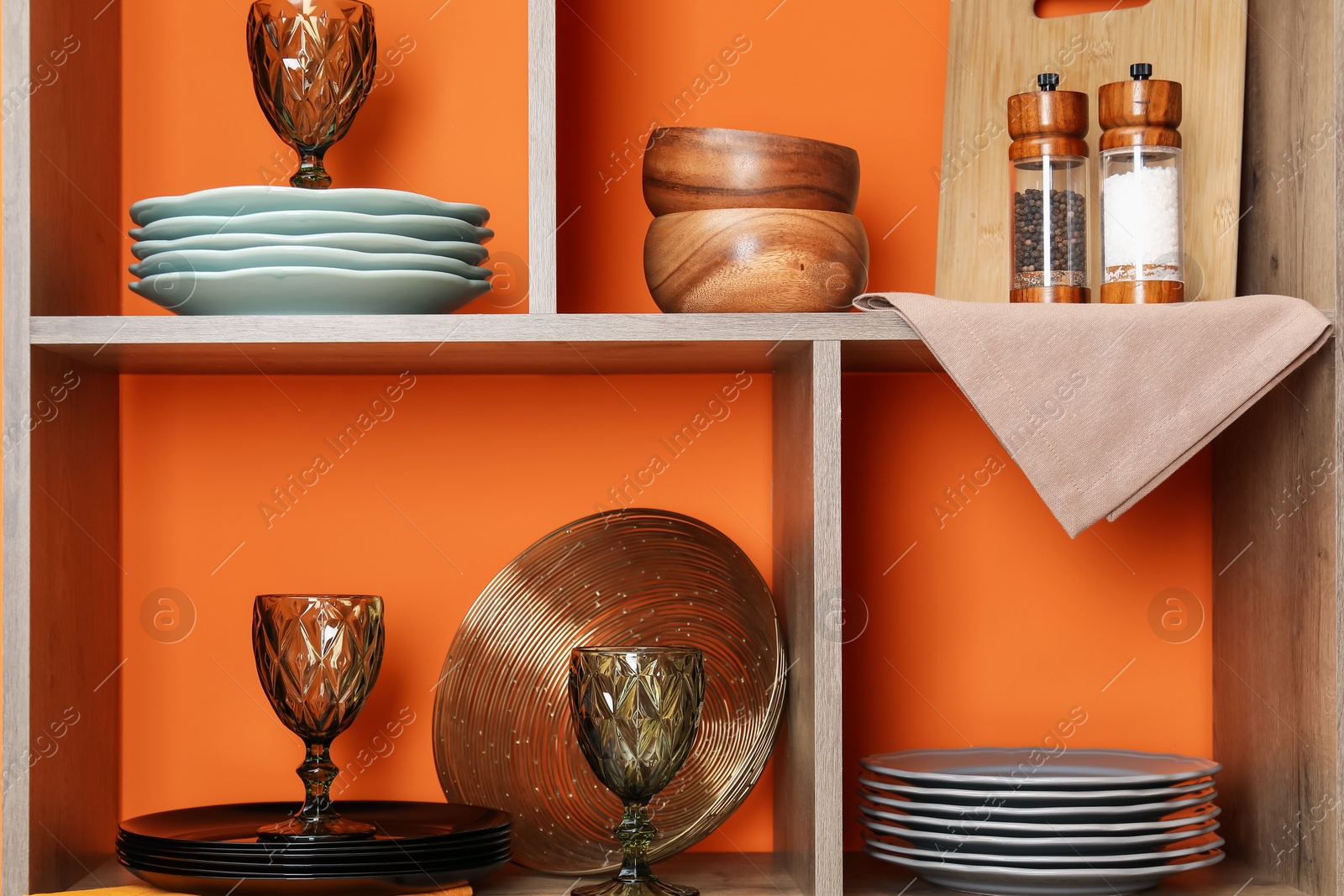 Photo of Shelving unit with different dishware and glasses near orange wall indoors