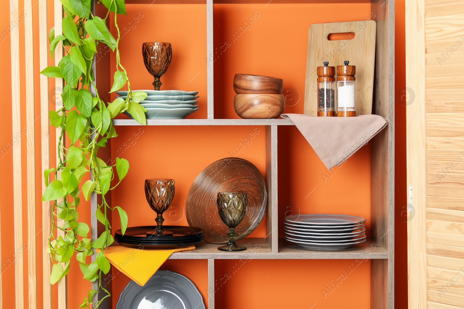 Photo of Shelving unit with different dishware and glasses near orange wall indoors