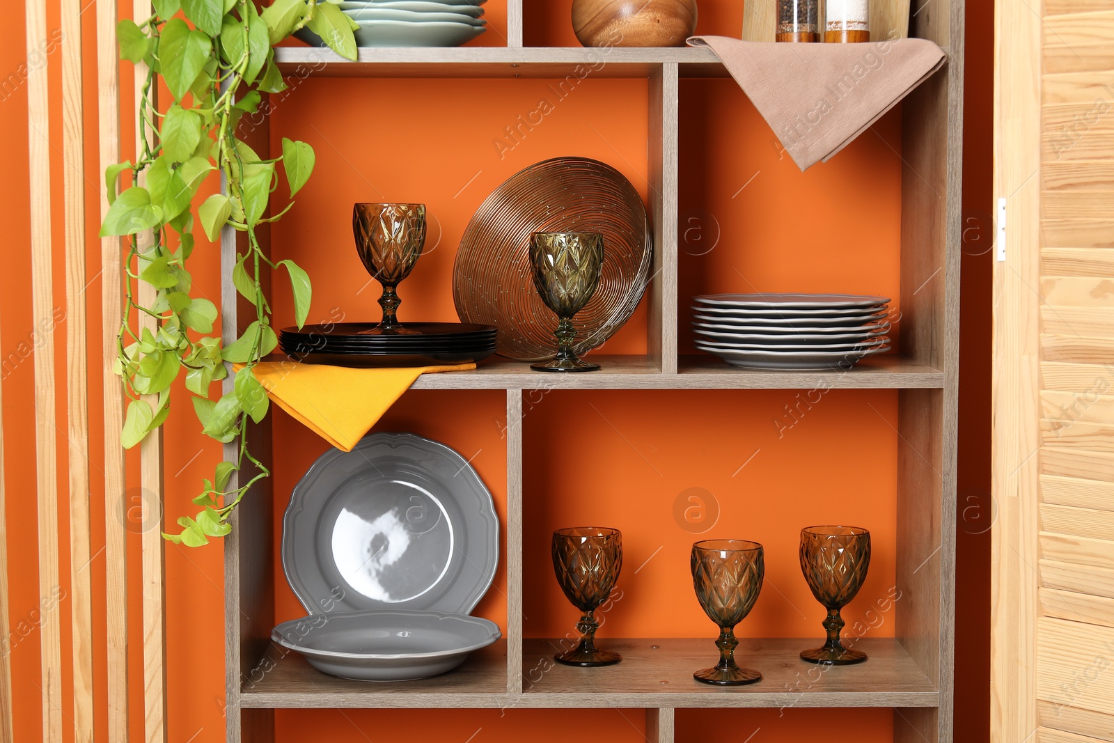 Photo of Shelving unit with different dishware and glasses near orange wall indoors