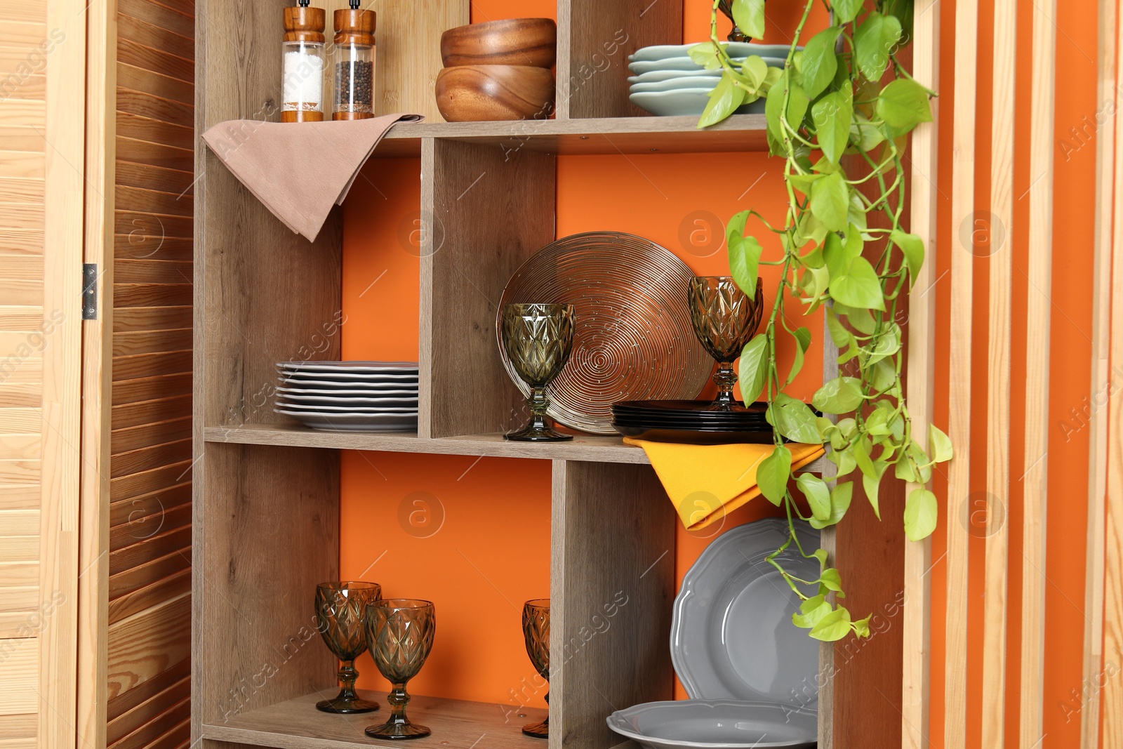 Photo of Shelving unit with different dishware and glasses near orange wall indoors