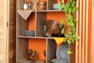 Photo of Shelving unit with different dishware and glasses near orange wall indoors