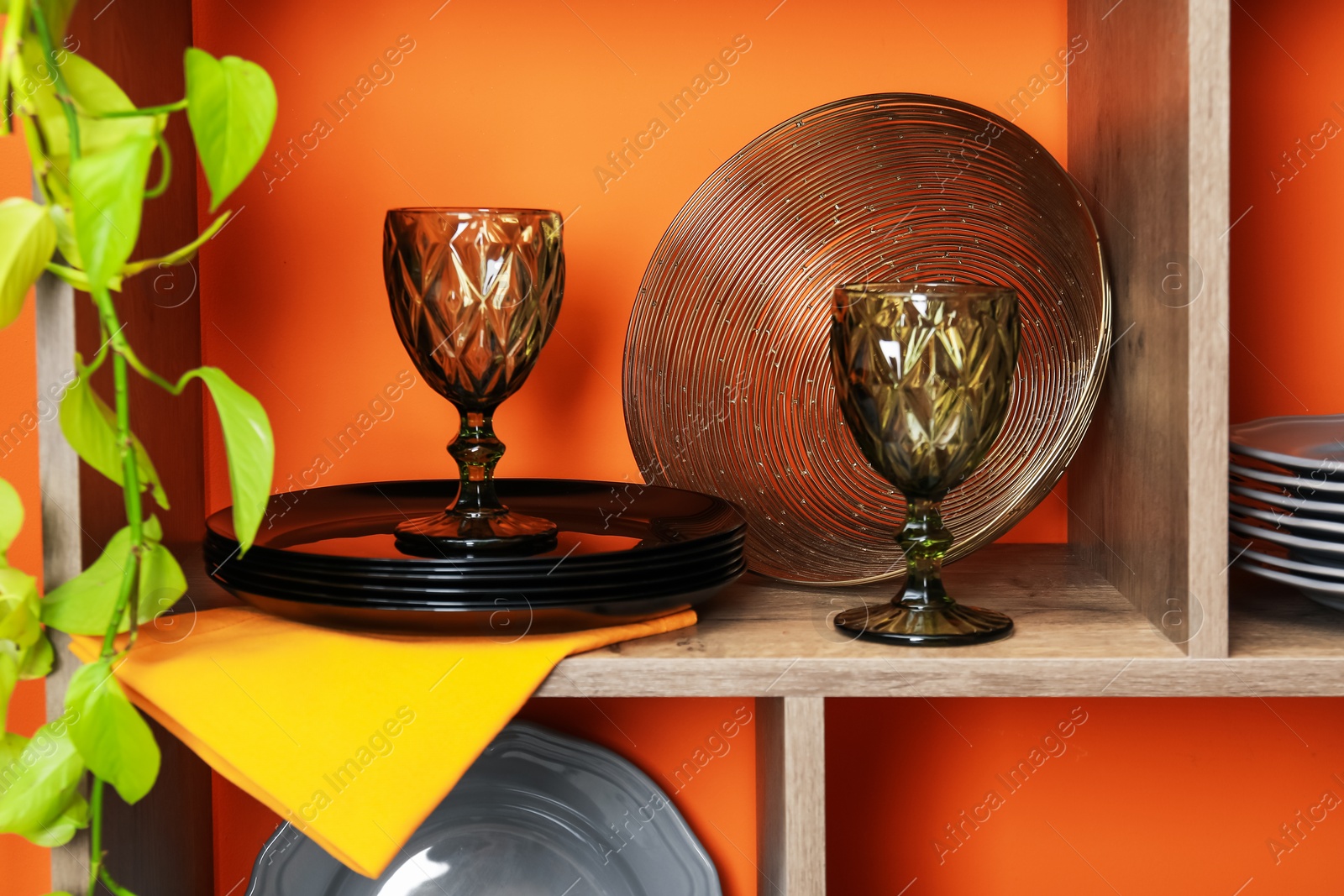 Photo of Shelving unit with different dishware and glasses near orange wall indoors, closeup