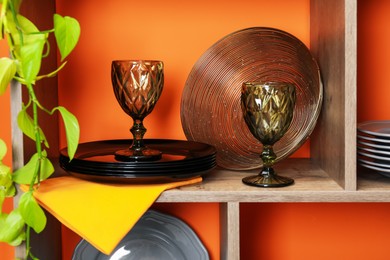 Photo of Shelving unit with different dishware and glasses near orange wall indoors, closeup