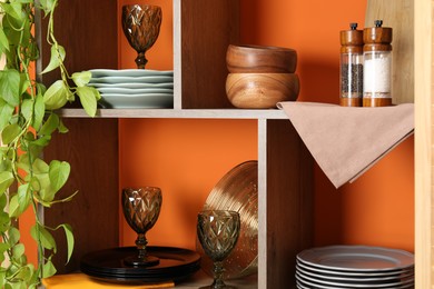 Photo of Shelving unit with different dishware and glasses near orange wall indoors