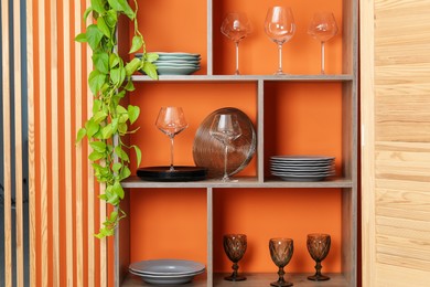 Photo of Shelving unit with different dishware and glasses near orange wall indoors