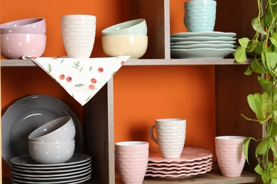 Photo of Shelving unit with different dishware and cups near orange wall indoors