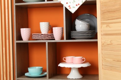 Photo of Shelving unit with different dishware and cups near orange wall indoors