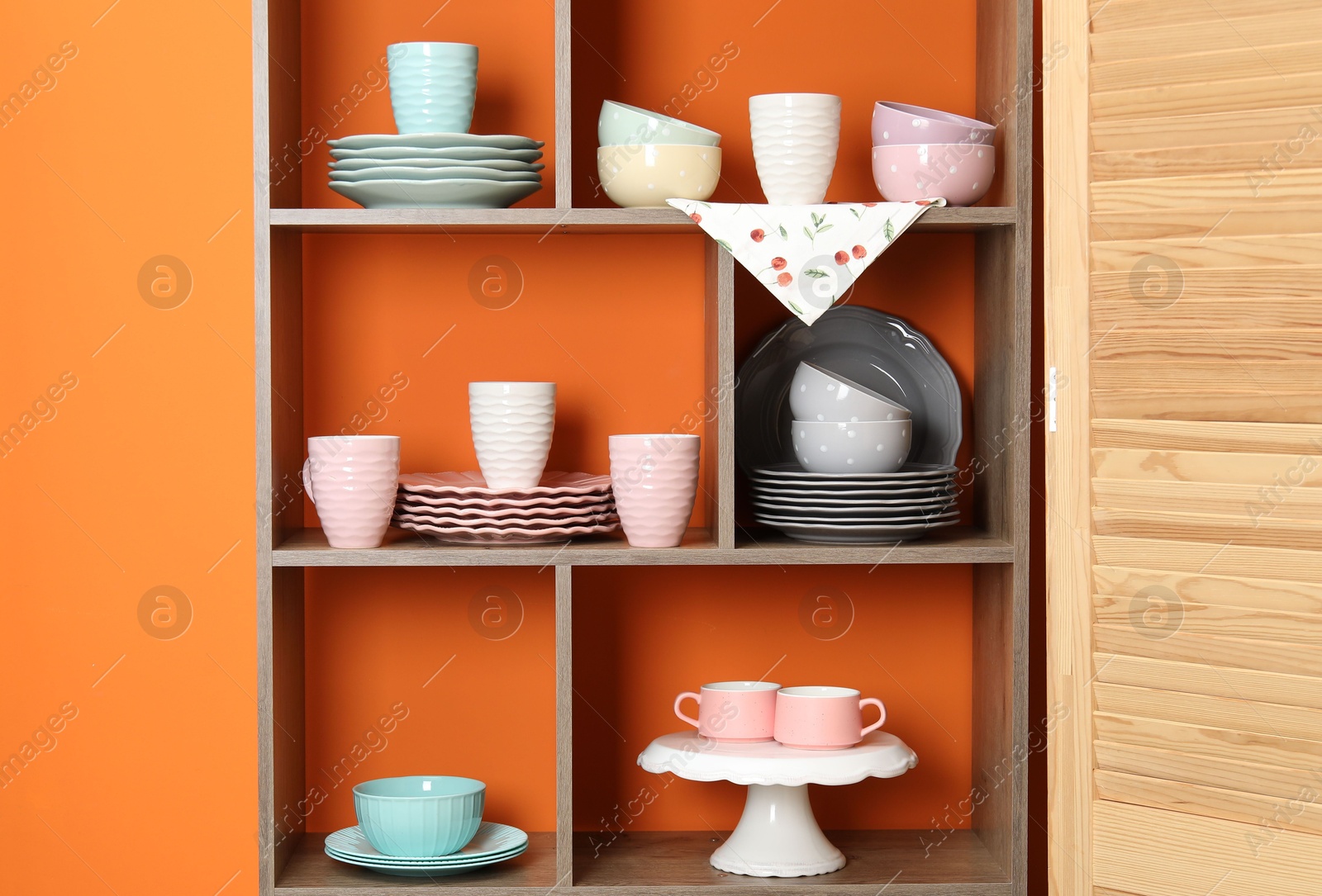Photo of Shelving unit with different dishware and cups near orange wall indoors