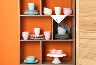 Photo of Shelving unit with different dishware and cups near orange wall indoors