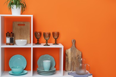 Photo of Shelving unit with different dishware, cups and floral decor near orange wall indoors