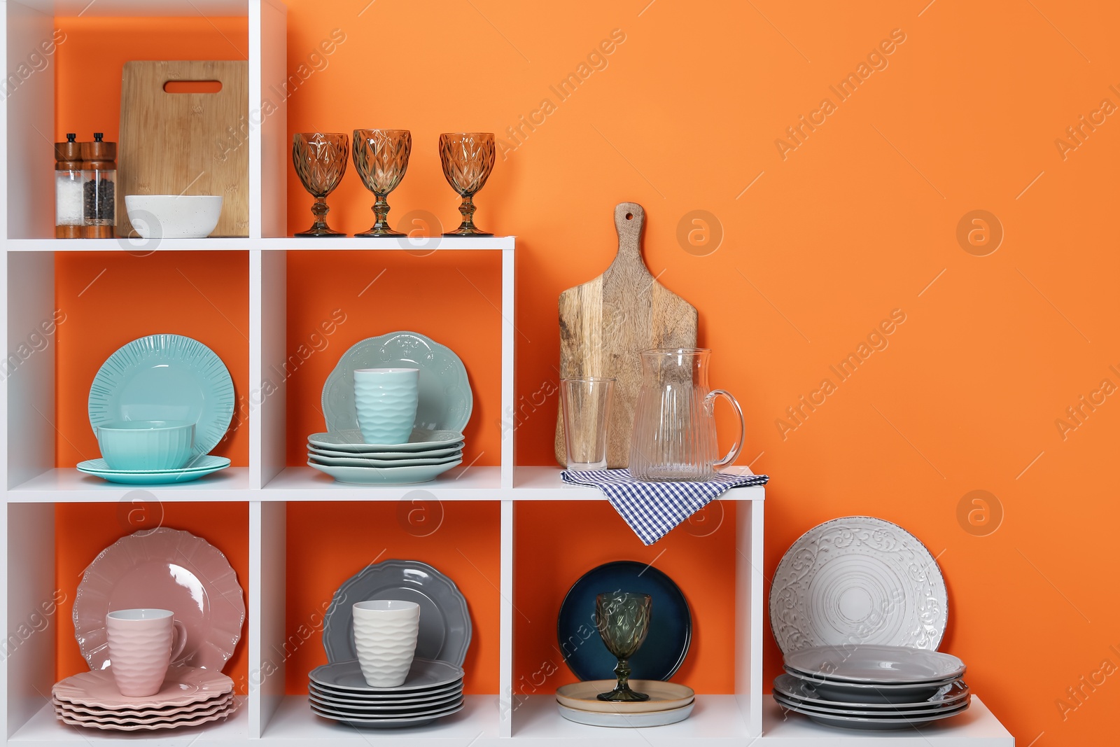 Photo of Shelving unit with different dishware, cups and glasses near orange wall indoors