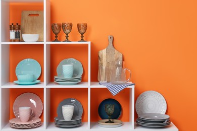 Photo of Shelving unit with different dishware, cups and glasses near orange wall indoors