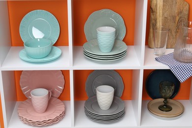 Photo of Shelving unit with different dishware, cups and glasses near orange wall indoors