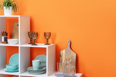 Photo of Shelving unit with different dishware, cups, glasses and floral decor near orange wall indoors