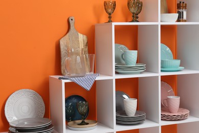 Photo of Shelving unit with different dishware, cups and glasses near orange wall indoors