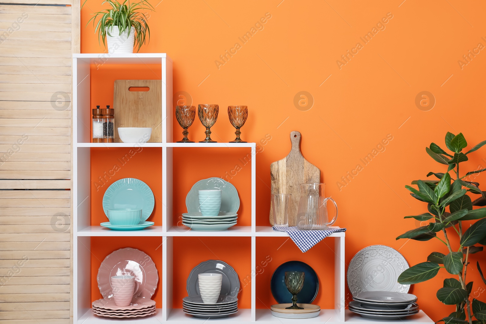Photo of Shelving unit with different dishware, cups, glasses and floral decor near orange wall indoors