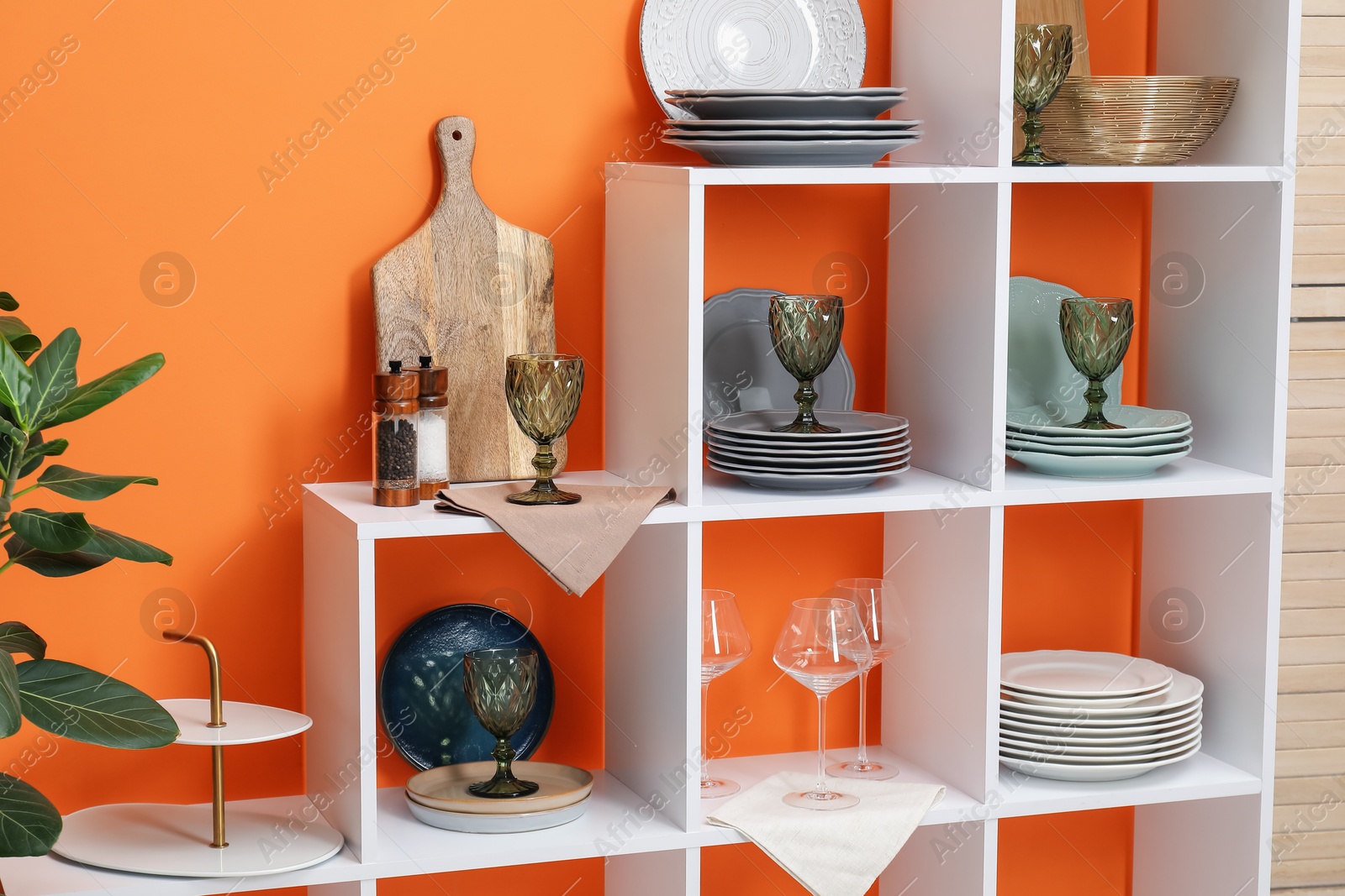 Photo of Shelving unit with different dishware and glasses near orange wall indoors