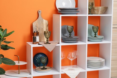 Photo of Shelving unit with different dishware and glasses near orange wall indoors