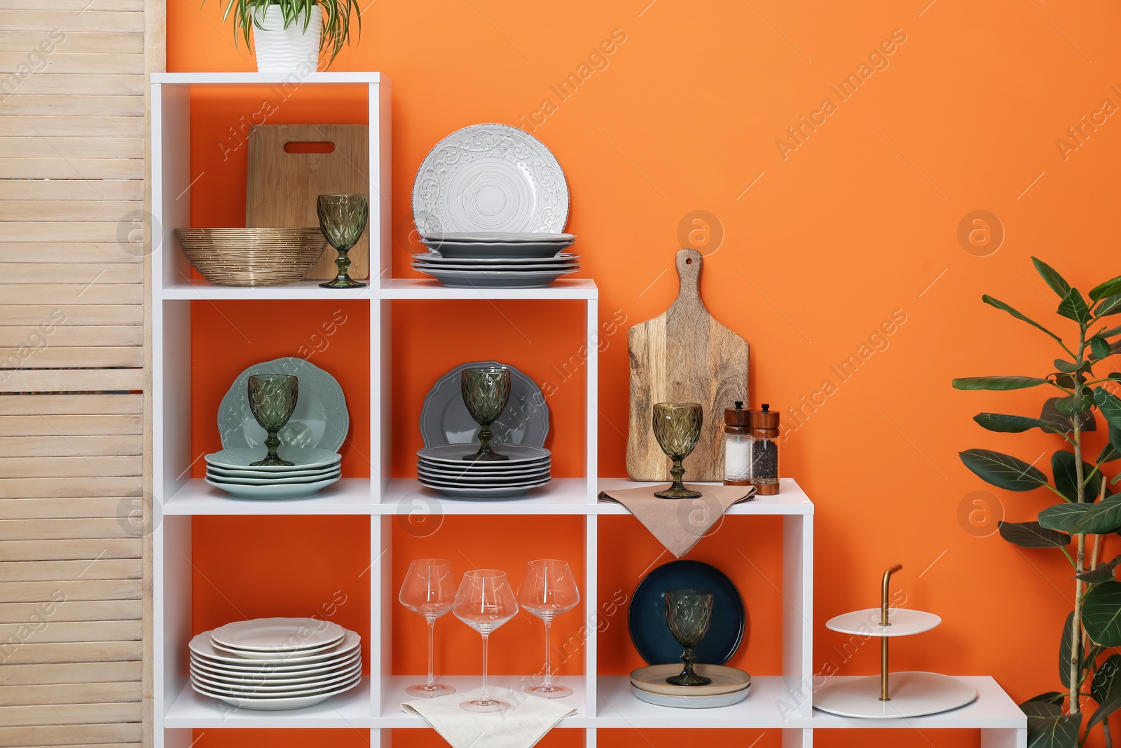 Photo of Shelving unit with different dishware, glasses and floral decor near orange wall indoors