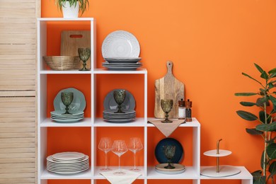 Photo of Shelving unit with different dishware, glasses and floral decor near orange wall indoors