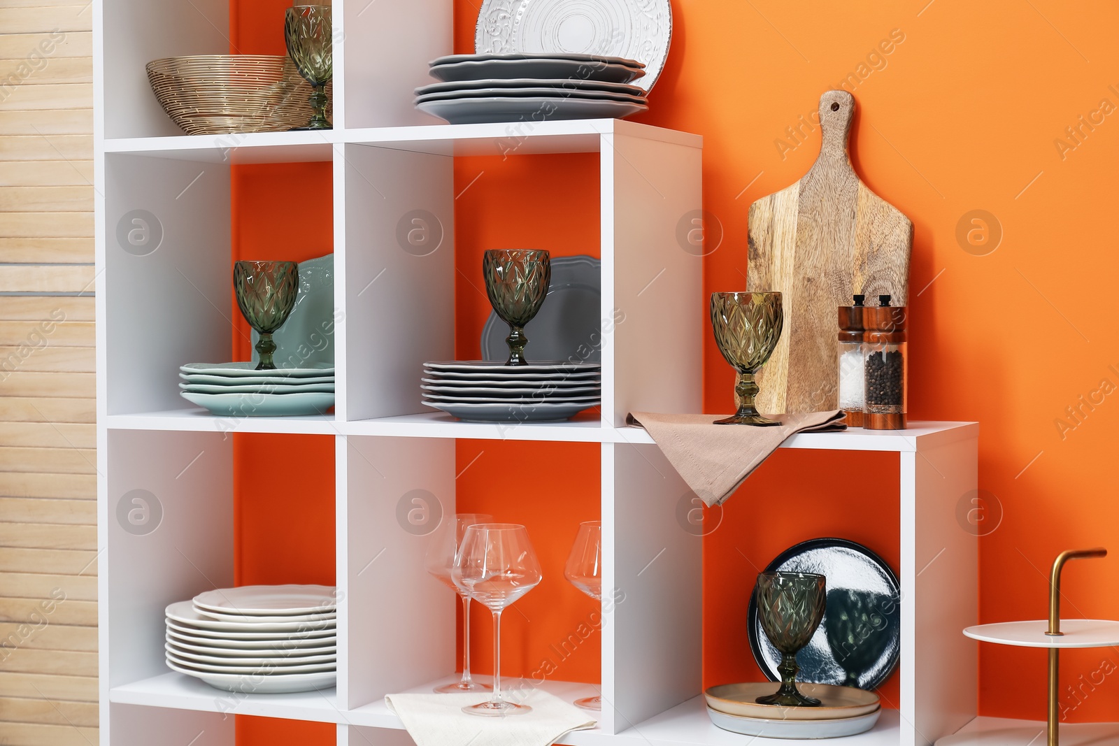 Photo of Shelving unit with different dishware and glasses near orange wall indoors