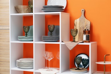 Photo of Shelving unit with different dishware and glasses near orange wall indoors