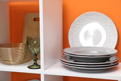 Photo of Different dishware and glass on shelves near orange wall indoors, closeup