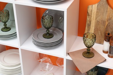 Photo of Shelving unit with different dishware and glasses near orange wall indoors, closeup