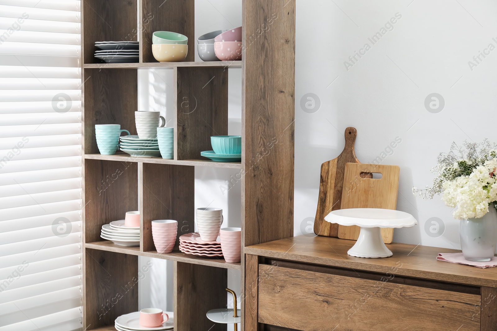 Photo of Shelving unit with different dishware, cups and floral decor near white wall indoors